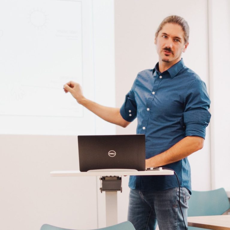 Mann in blauem Hemd präsentiert an einem Laptop vor einer weißen Wand.