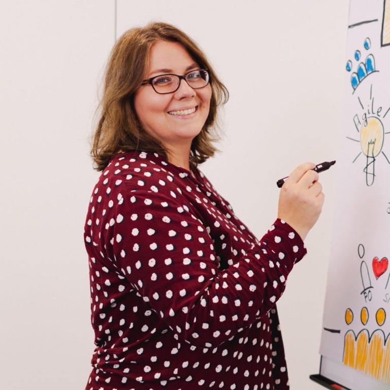 Frau mit Brille und gepunktetem Hemd, die an einem Flipchart zeichnet.